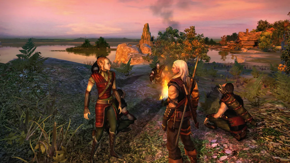 Geralt z towarzyszami przy ognisku nad jeziorem o zachodzie słońca w grze Wiedźmin z 2007 roku